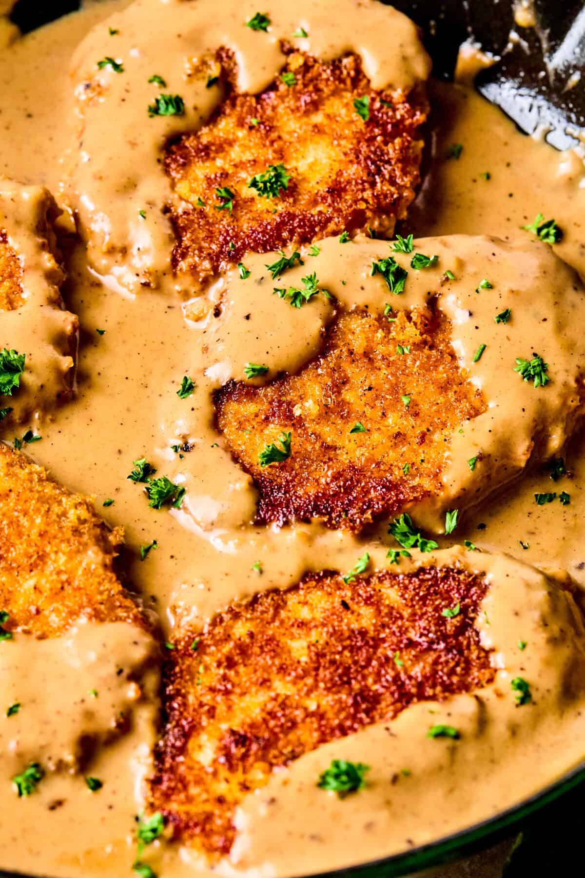 Crispy breaded pork chops and gravy, covered in creamy brown sauce and garnished with chopped parsley, served sizzling in a skillet.