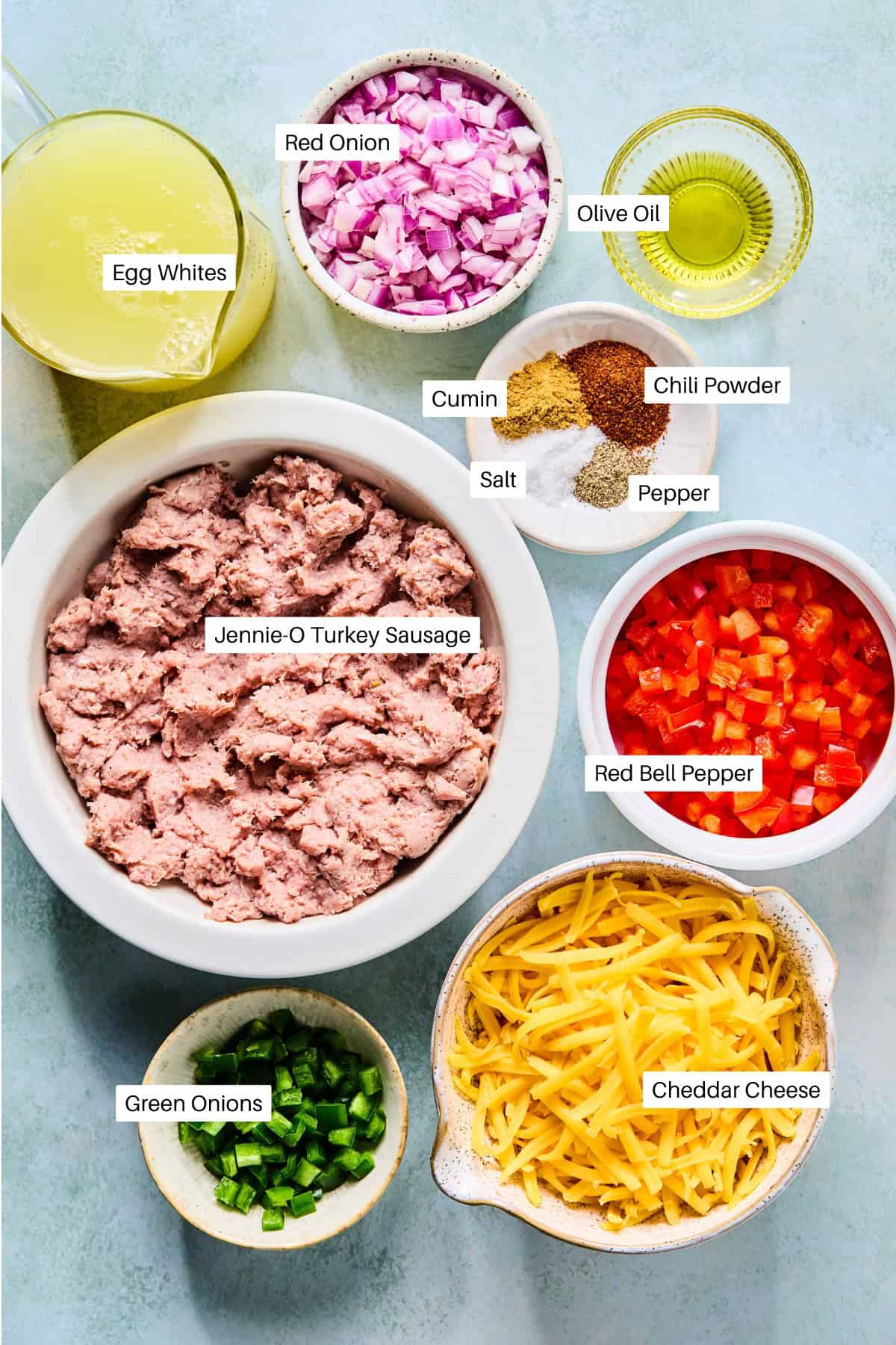 Bowls of labeled ingredients for a southwest frittata on a light surface: egg whites, red onion, olive oil, cumin, chili powder, salt, pepper, red bell pepper, Jennie-O turkey sausage, green onions, and cheddar cheese.