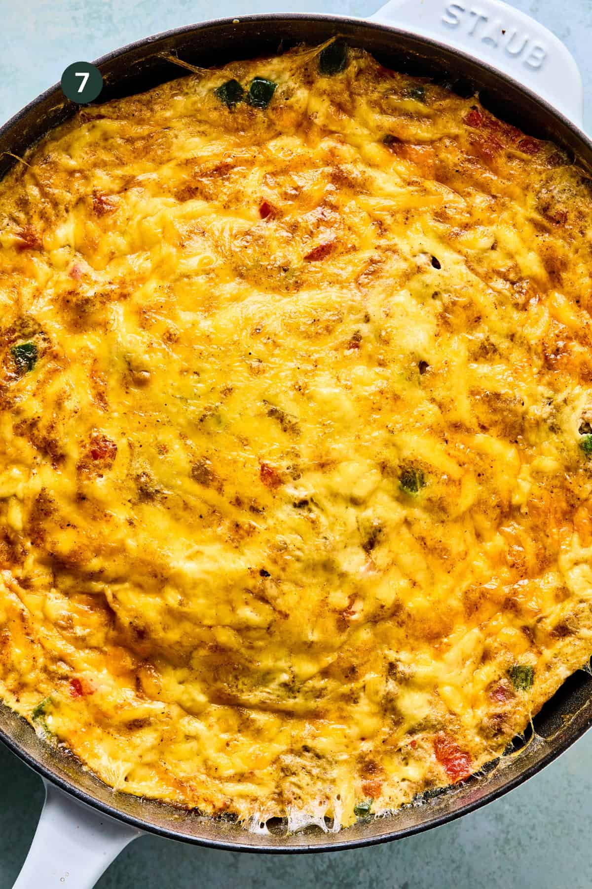 A close-up of a golden-brown southwest frittata in a round white skillet, featuring melted cheese and bits of vegetables on top.