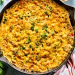 A cast iron skillet filled with a golden, cheesy southwest frittata casserole, garnished with chopped herbs. Surrounding the skillet are small bowls of salsa, sour cream, and fresh herbs, with a red-and-white striped towel nearby.