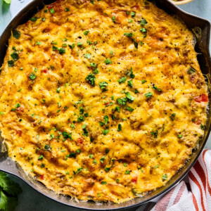 A cast iron skillet filled with a golden, cheesy southwest frittata casserole, garnished with chopped herbs. Surrounding the skillet are small bowls of salsa, sour cream, and fresh herbs, with a red-and-white striped towel nearby.