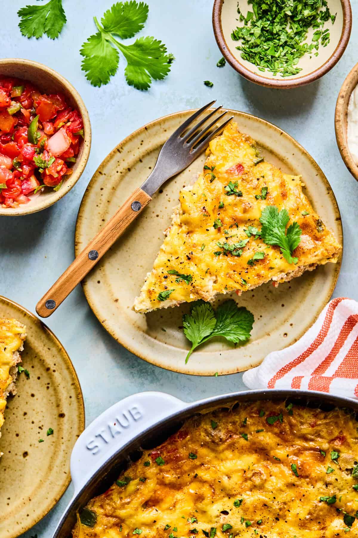 A slice of cheesy southwest frittata garnished with cilantro sits on a tan plate with a fork. Surrounding the plate are bowls of chopped herbs, salsa, and a casserole dish filled with more frittata.