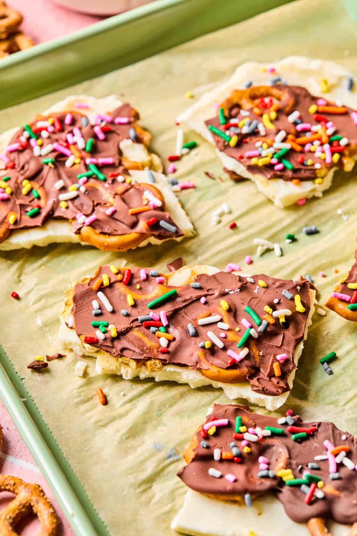 Rectangular pieces of white crackers topped with melted chocolate and colorful sprinkles are arranged on a parchment-lined tray, resembling a playful twist on protein frozen yogurt bark. Whole pretzels and crumbs add extra crunch around the treats.