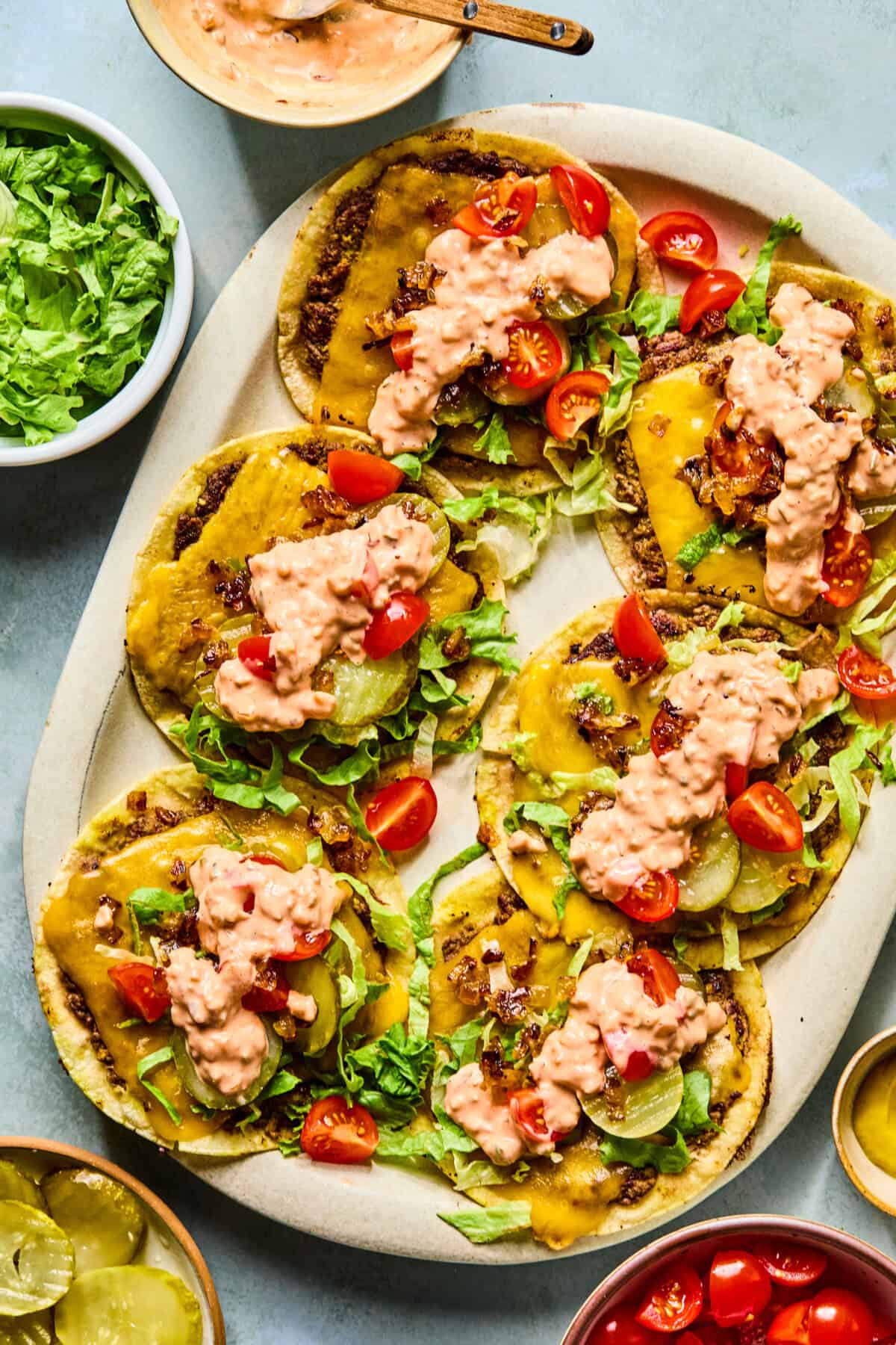A platter of open-faced smash tacos topped with shredded lettuce, sliced pickles, diced tomatoes, melted cheese, and a creamy pink sauce. Bowls of lettuce, pickles, cherry tomatoes, and sauce surround the tacos.