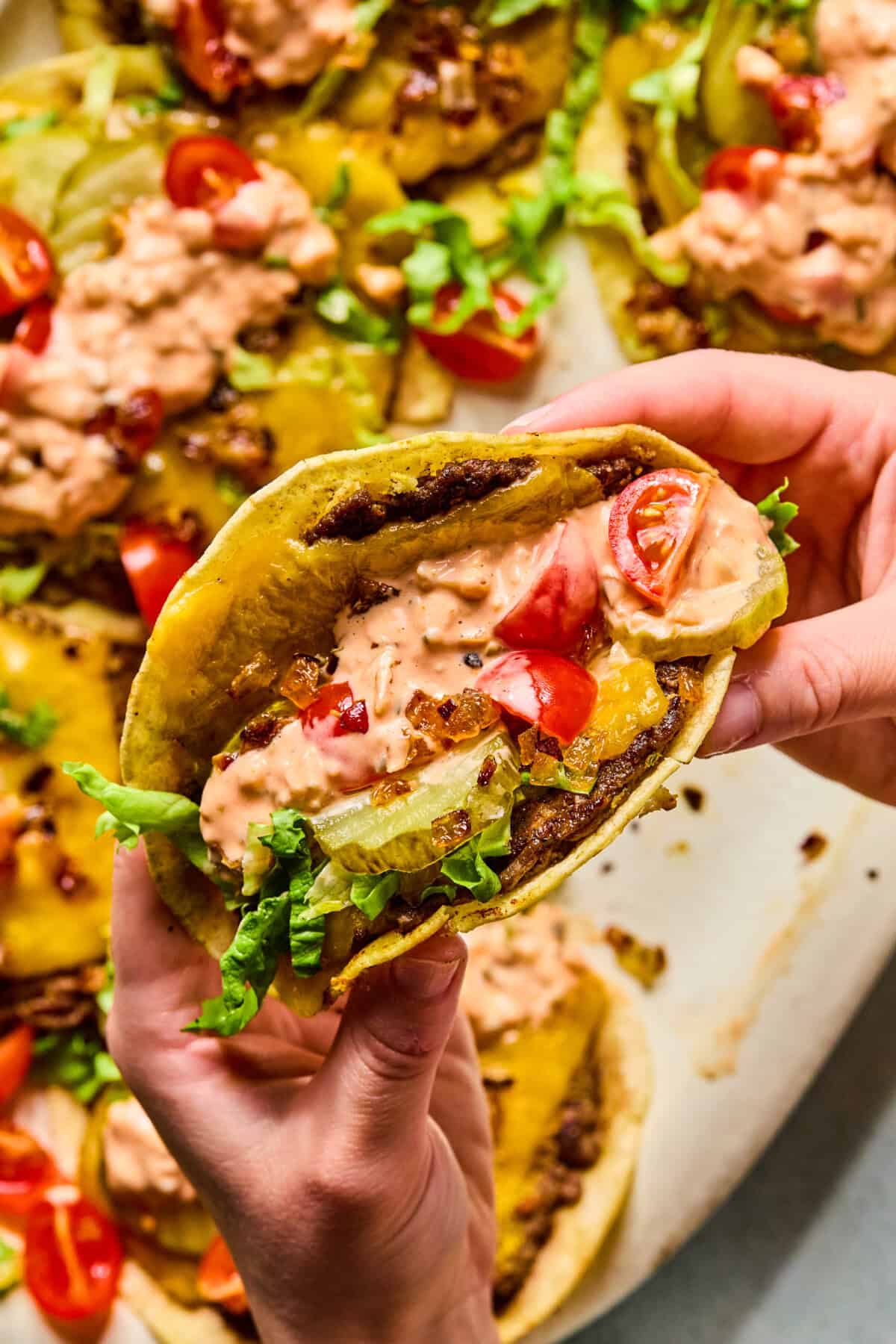 A close-up of hands holding a smash taco filled with lettuce, tomato, cheddar cheese, ground beef, pickles, and creamy sauce. More smash tacos with similar toppings are visible in the background.