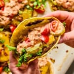 A close-up of hands holding a smash taco filled with lettuce, tomato, cheddar cheese, ground beef, pickles, and creamy sauce. More smash tacos with similar toppings are visible in the background.