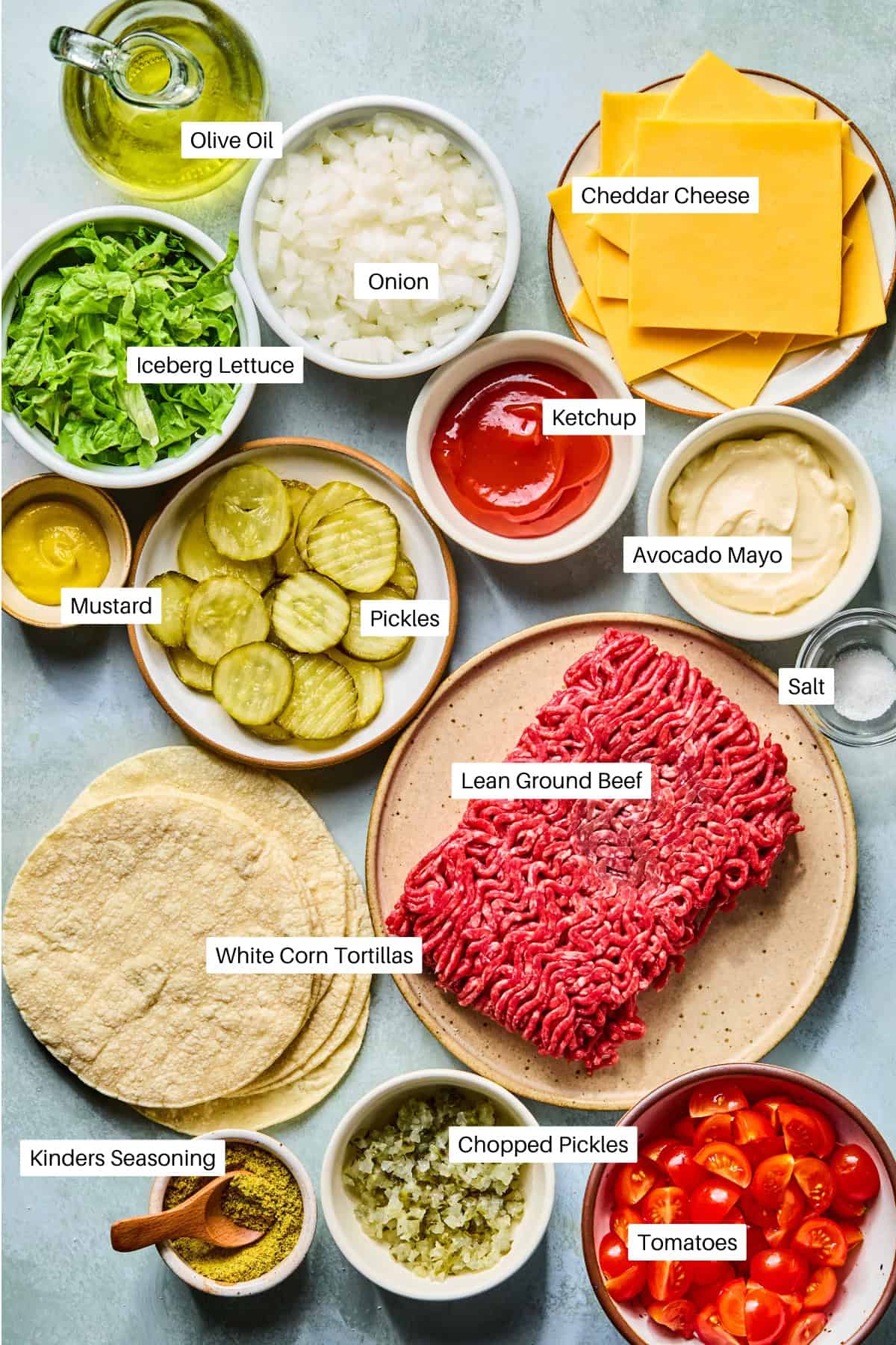 An overhead view of labeled ingredients for smash tacos, including lean ground beef, cheddar cheese, white corn tortillas, onion, iceberg lettuce, pickles, chopped pickles, tomatoes, ketchup, mustard, avocado mayo, salt, olive oil, and seasoning.