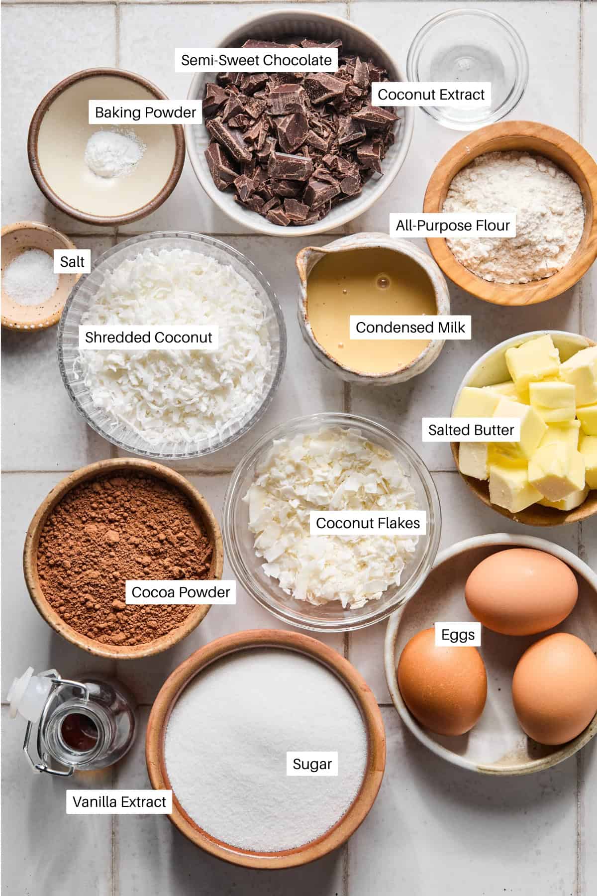 A top view of various baking ingredients for coconut brownies in bowls on a white surface, including eggs, butter, condensed milk, shredded coconut, coconut flakes, cocoa powder, semi-sweet chocolate, flour, sugar, extracts, salt, and baking powder.