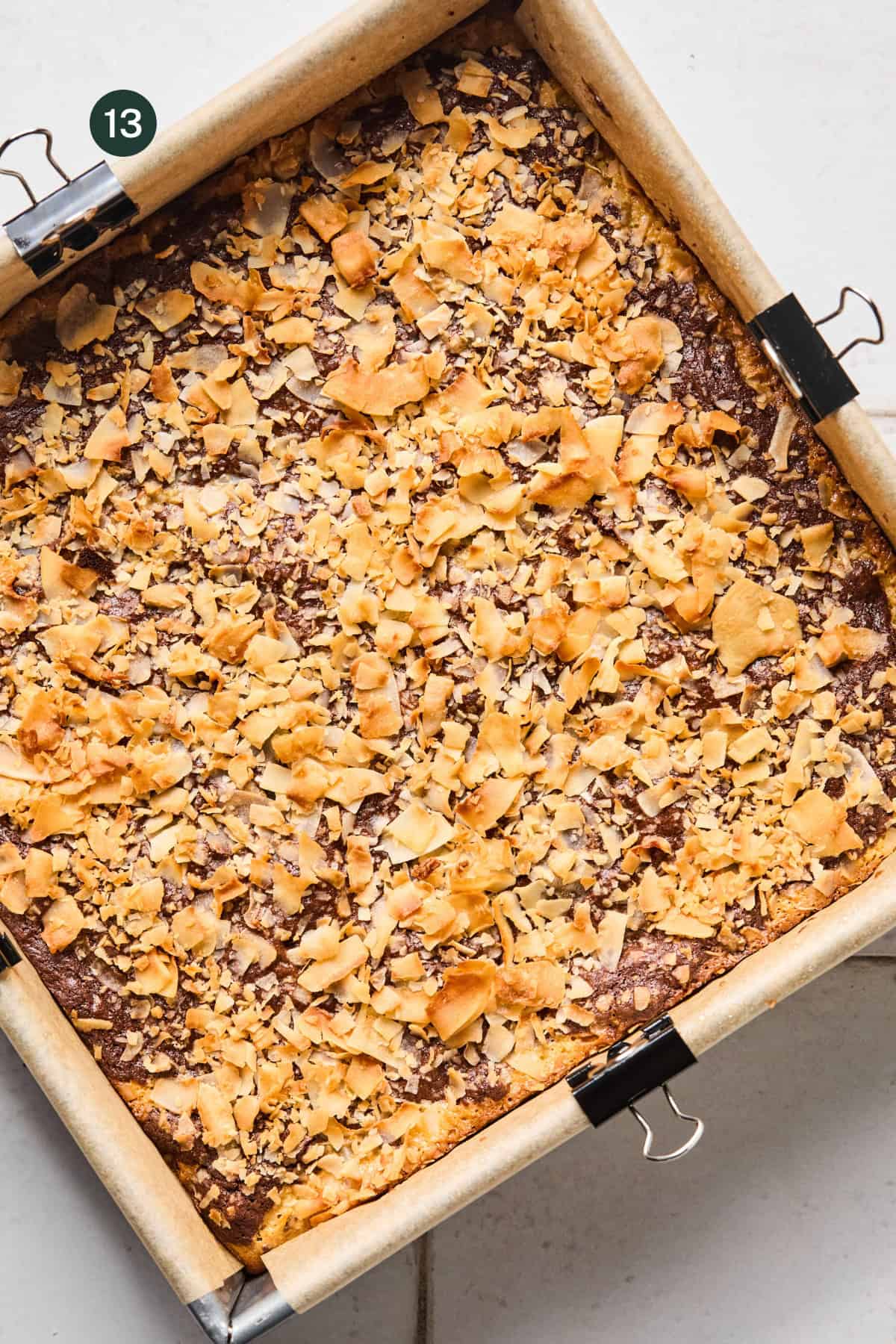 A square baking pan lined with parchment paper holds coconut brownies topped with golden, toasted coconut flakes. Two binder clips secure the parchment to the pan. The number 13 appears in the top left corner.