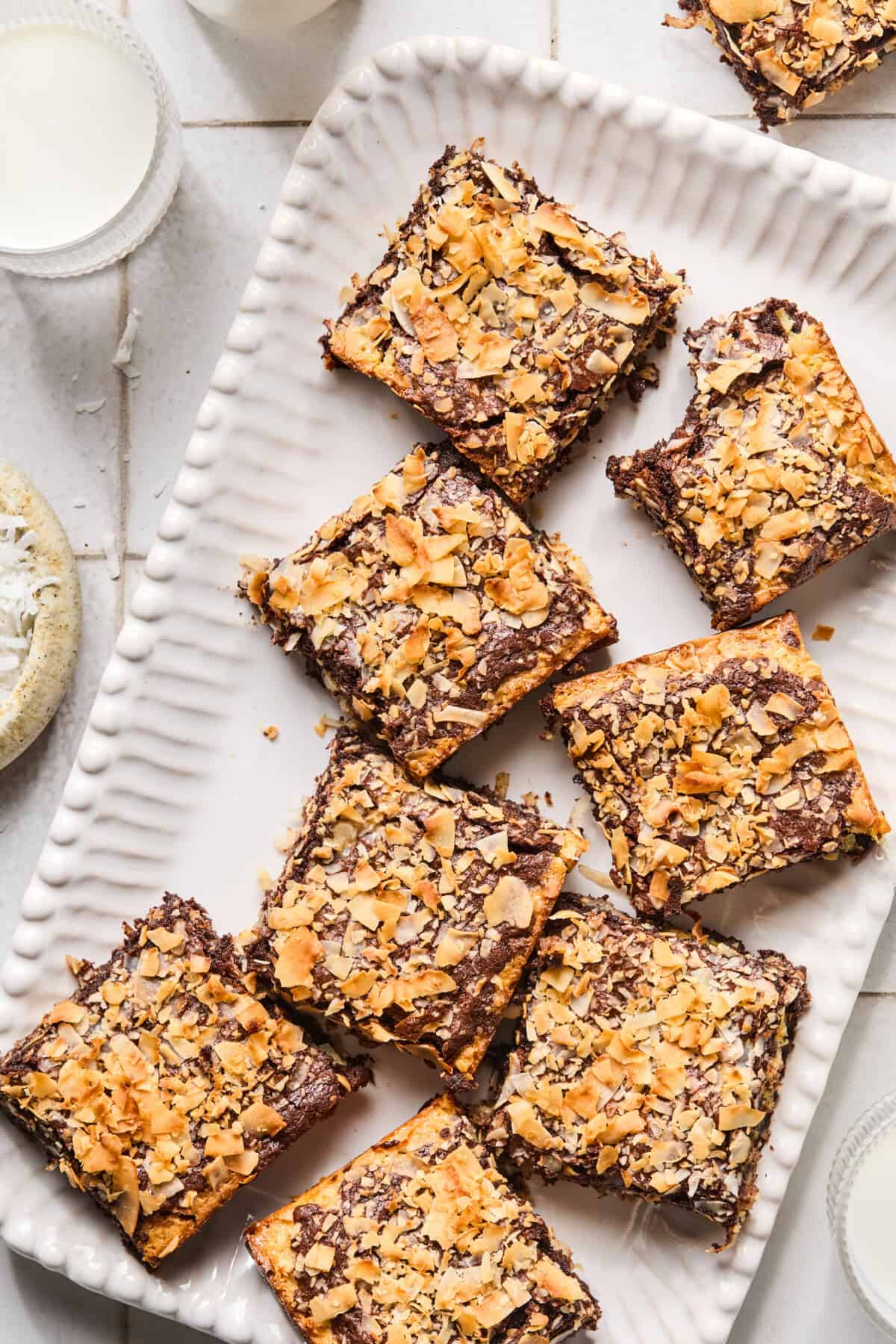 A white rectangular tray holds eight coconut brownies, some with bites taken out. The bars are topped with toasted coconut flakes. Glasses of milk and a bowl of shredded coconut are nearby.