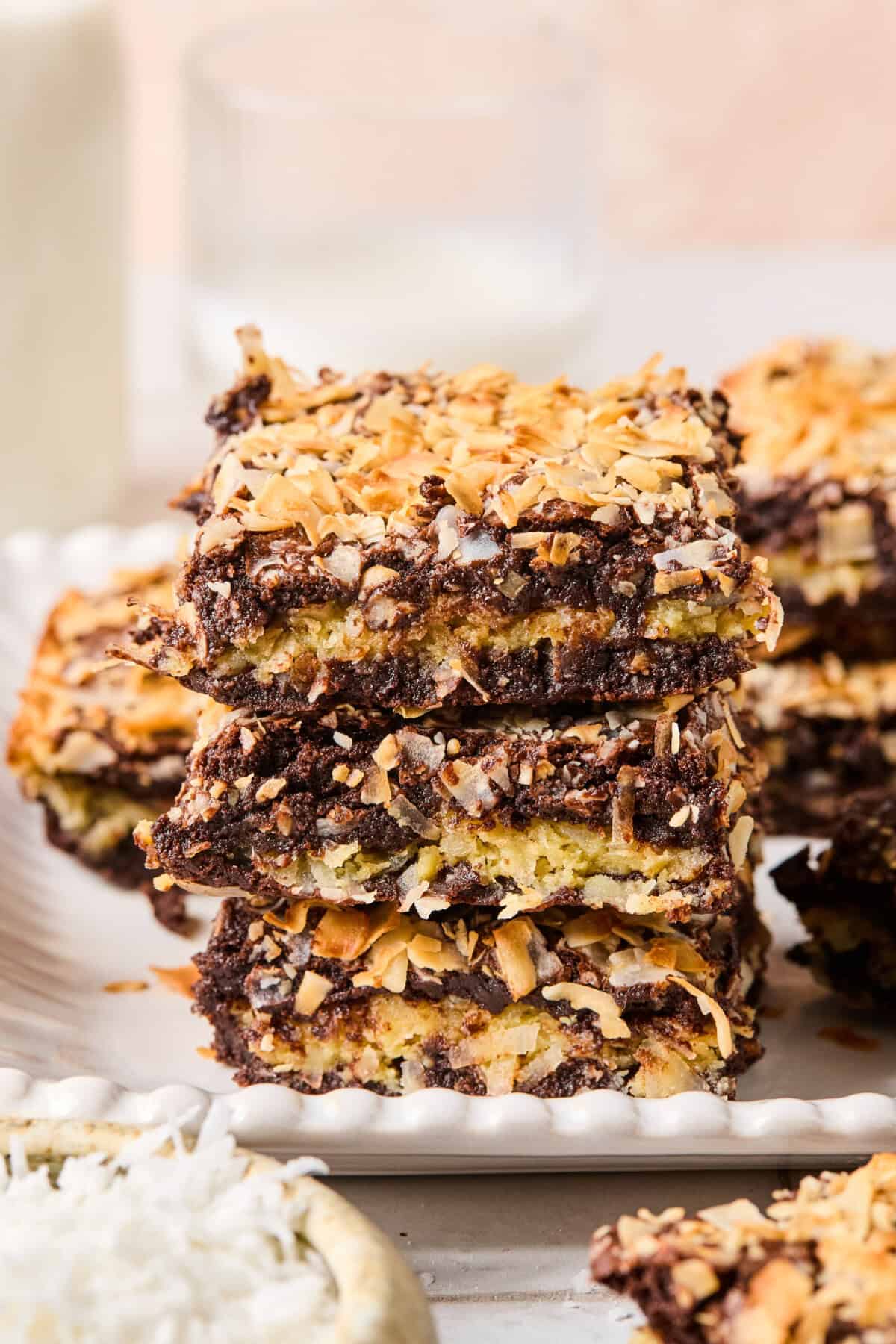 Three dessert bars are stacked on a plate, resembling coconut brownies with a chocolate and coconut topping and a golden, toasted appearance. More bars and a glass of milk are blurred in the background.