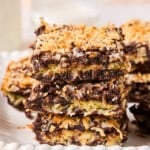 Three dessert bars are stacked on a plate, resembling coconut brownies with a chocolate and coconut topping and a golden, toasted appearance. More bars and a glass of milk are blurred in the background.