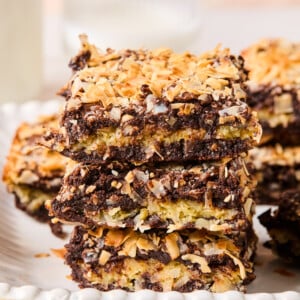 Three dessert bars are stacked on a plate, resembling coconut brownies with a chocolate and coconut topping and a golden, toasted appearance. More bars and a glass of milk are blurred in the background.