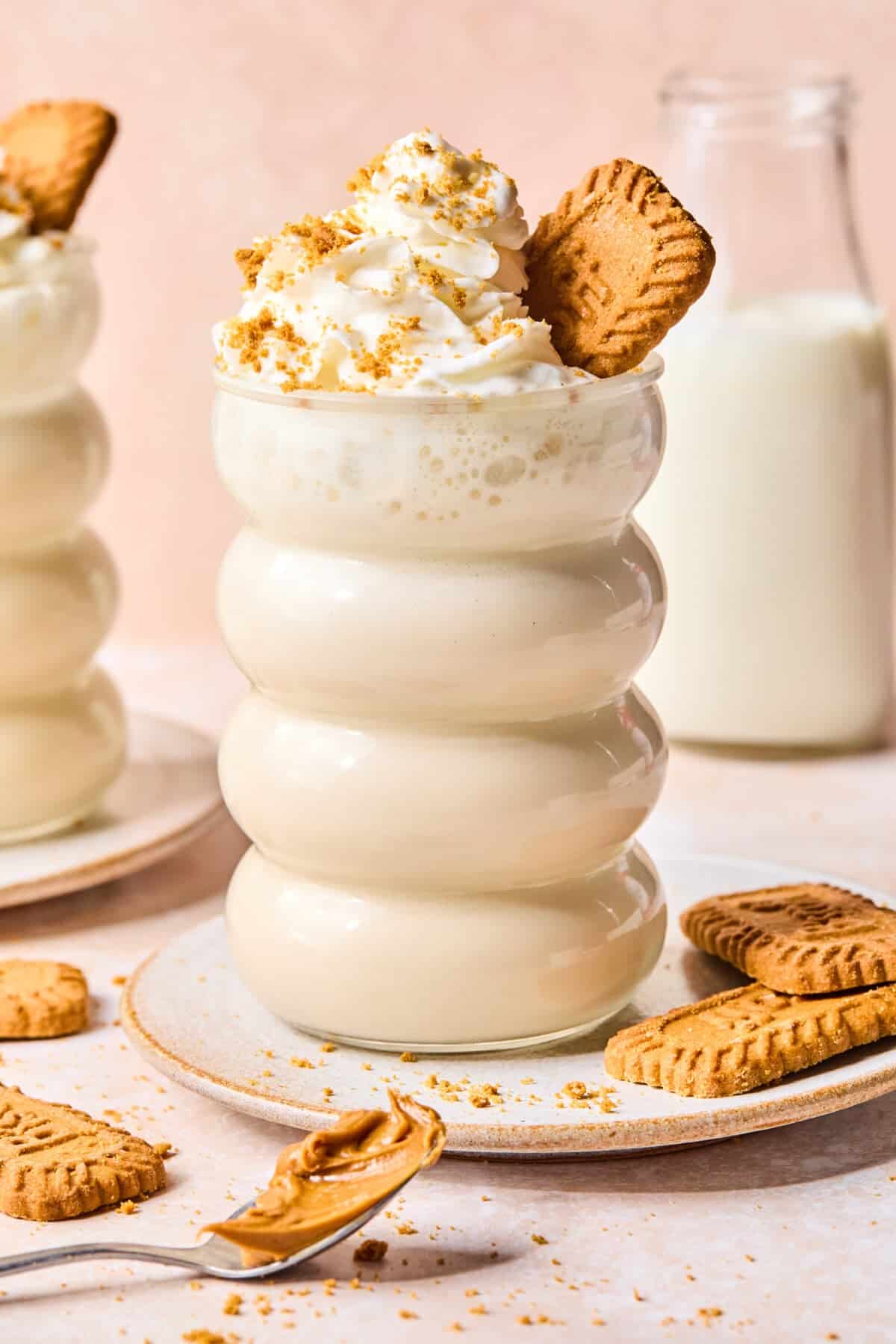 A creamy biscoff protein smoothie in a wavy glass, topped with whipped cream, cookie crumbs, and a whole cookie. A bottle of milk, extra cookies, and a spoon with cookie spread are nearby on a light surface.
