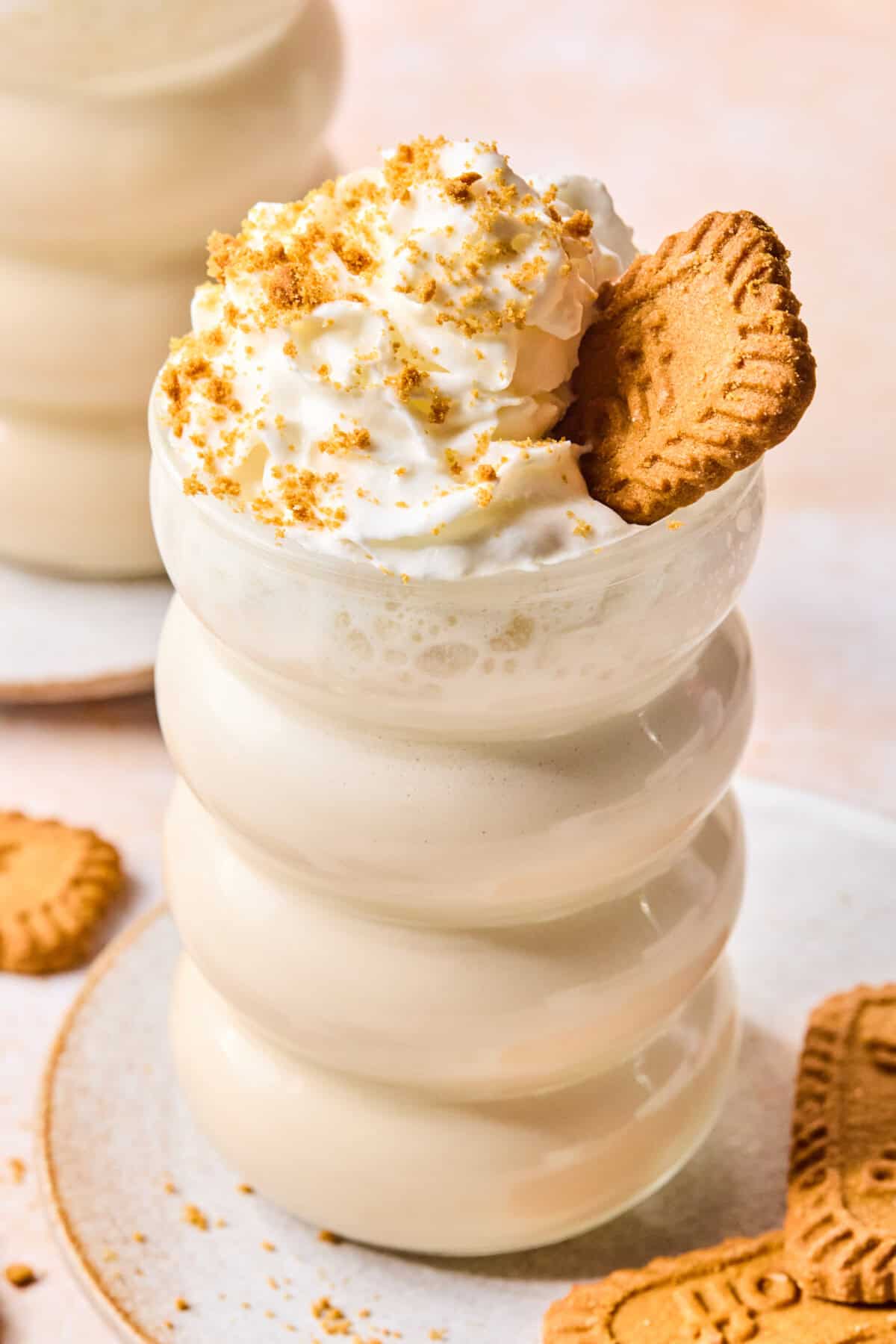A creamy biscoff protein smoothie topped with whipped cream, cookie crumbs, and a whole cookie sits in a wavy glass on a plate with more cookies nearby.