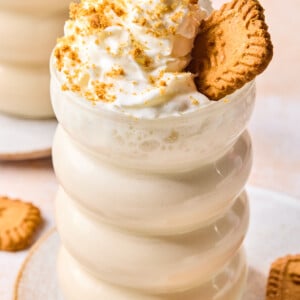 A creamy biscoff protein smoothie topped with whipped cream, cookie crumbs, and a whole cookie sits in a wavy glass on a plate with more cookies nearby.