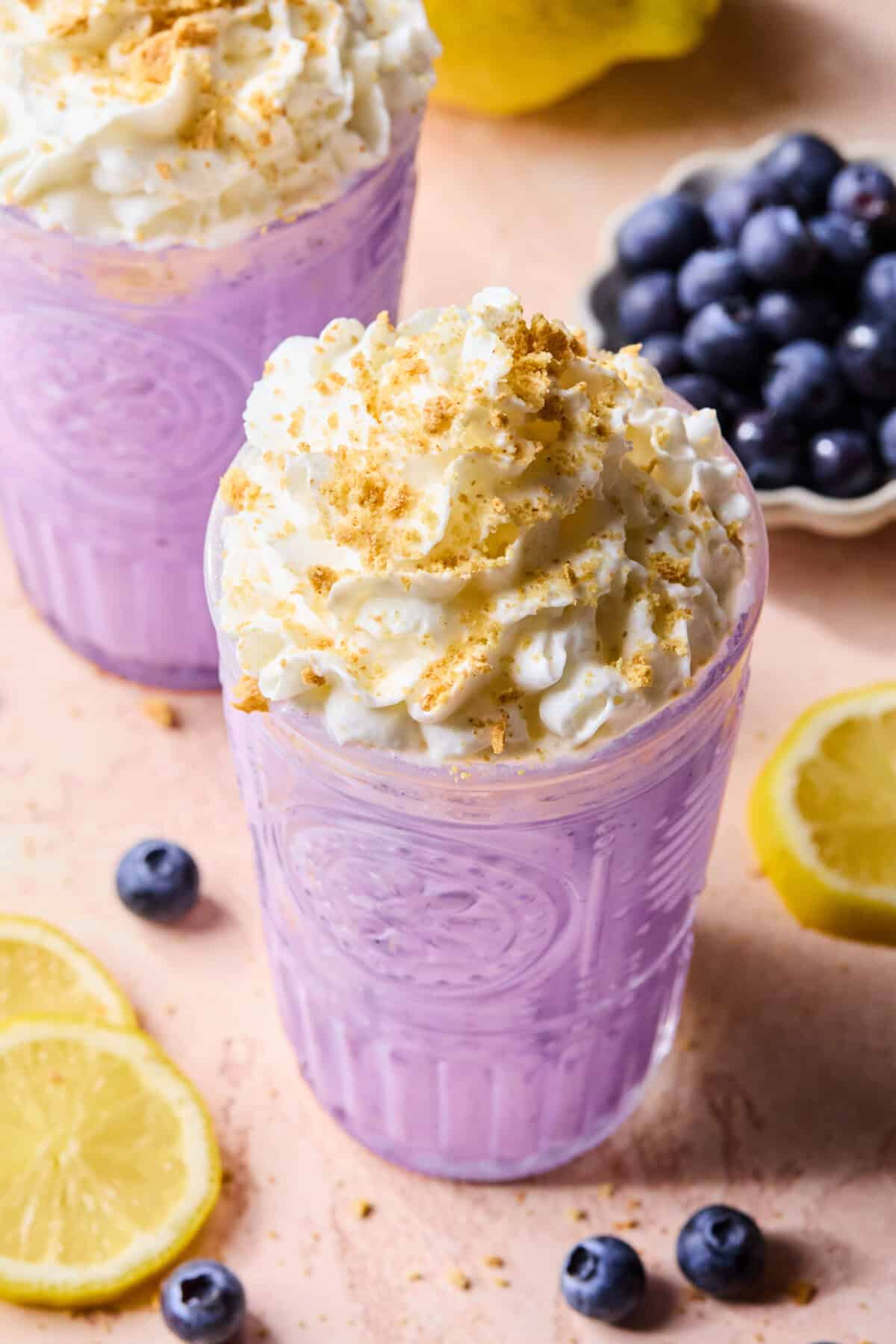 Two glasses of rich blueberry protein smoothie milkshake are topped with whipped cream and cookie crumbles. Fresh blueberries and lemon slices are scattered on a light surface, with a bowl of blueberries and a lemon in the background.