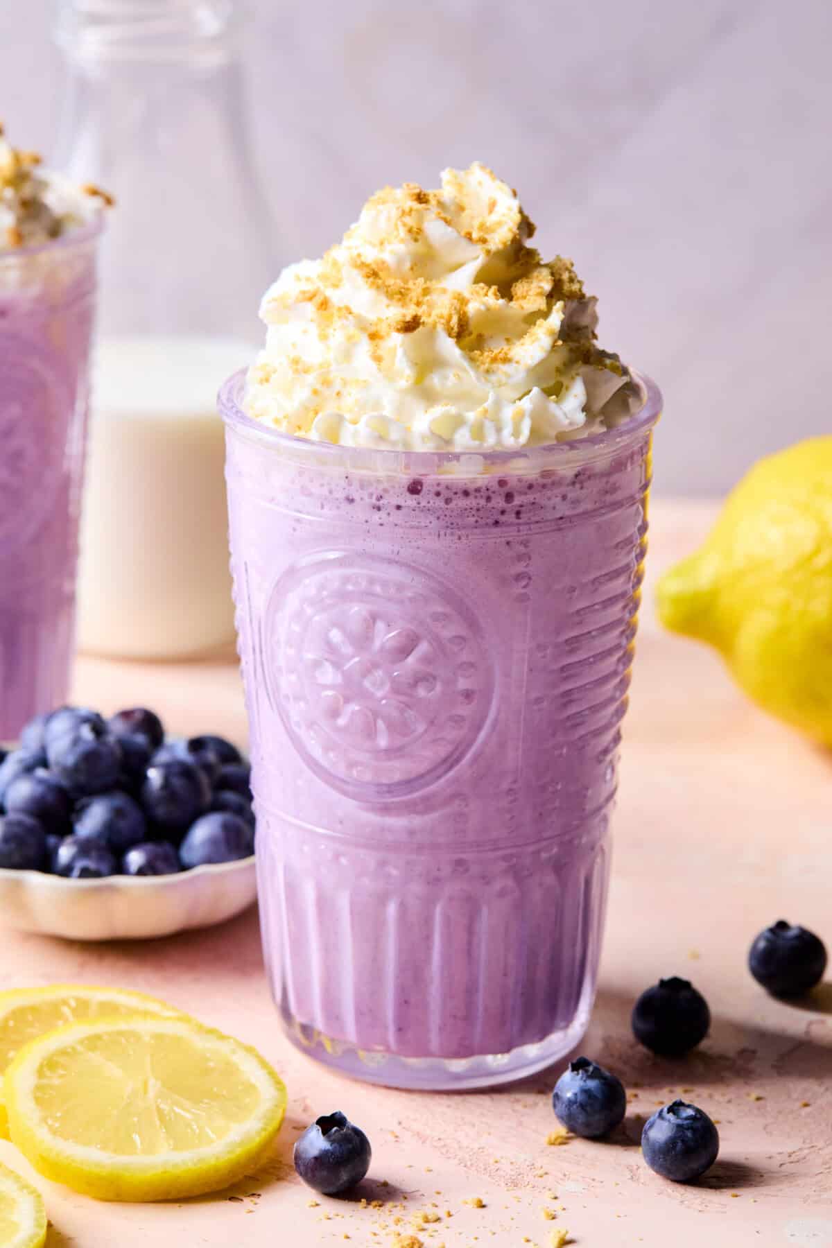 A purple blueberry protein smoothie in a clear glass topped with whipped cream and crumbs, surrounded by fresh blueberries and lemon slices, with a milk bottle and lemon in the background.