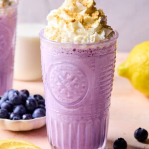 A purple blueberry protein smoothie in a clear glass topped with whipped cream and crumbs, surrounded by fresh blueberries and lemon slices, with a milk bottle and lemon in the background.