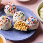 Four white chocolate-coated protein balls topped with colorful sprinkles sit on a purple plate. One ball is cut open, revealing a creamy peanut butter filling. Bowls of sprinkles and white chocolate chips surround the plate.