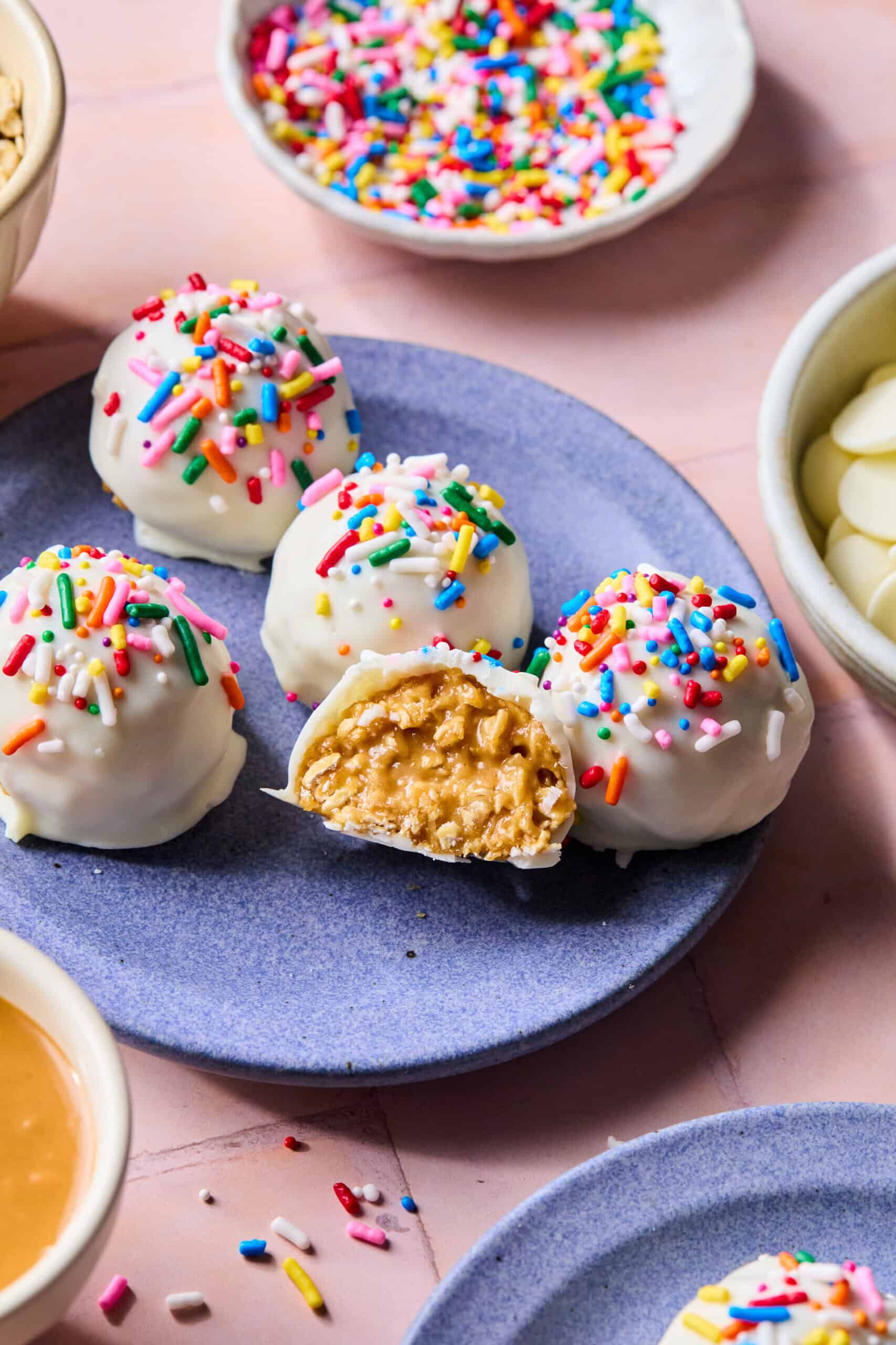 Four white chocolate-coated protein balls topped with colorful sprinkles sit on a purple plate. One ball is cut open, revealing a creamy peanut butter filling. Bowls of sprinkles and white chocolate chips surround the plate.