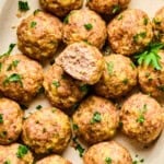 A platter of golden-brown baked turkey meatballs garnished with chopped parsley, with one meatball cut in half to reveal the juicy, tender inside.
