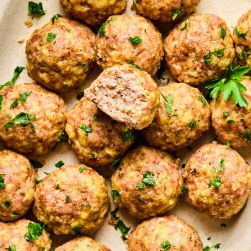A platter of golden-brown baked turkey meatballs garnished with chopped parsley, with one meatball cut in half to reveal the juicy, tender inside.