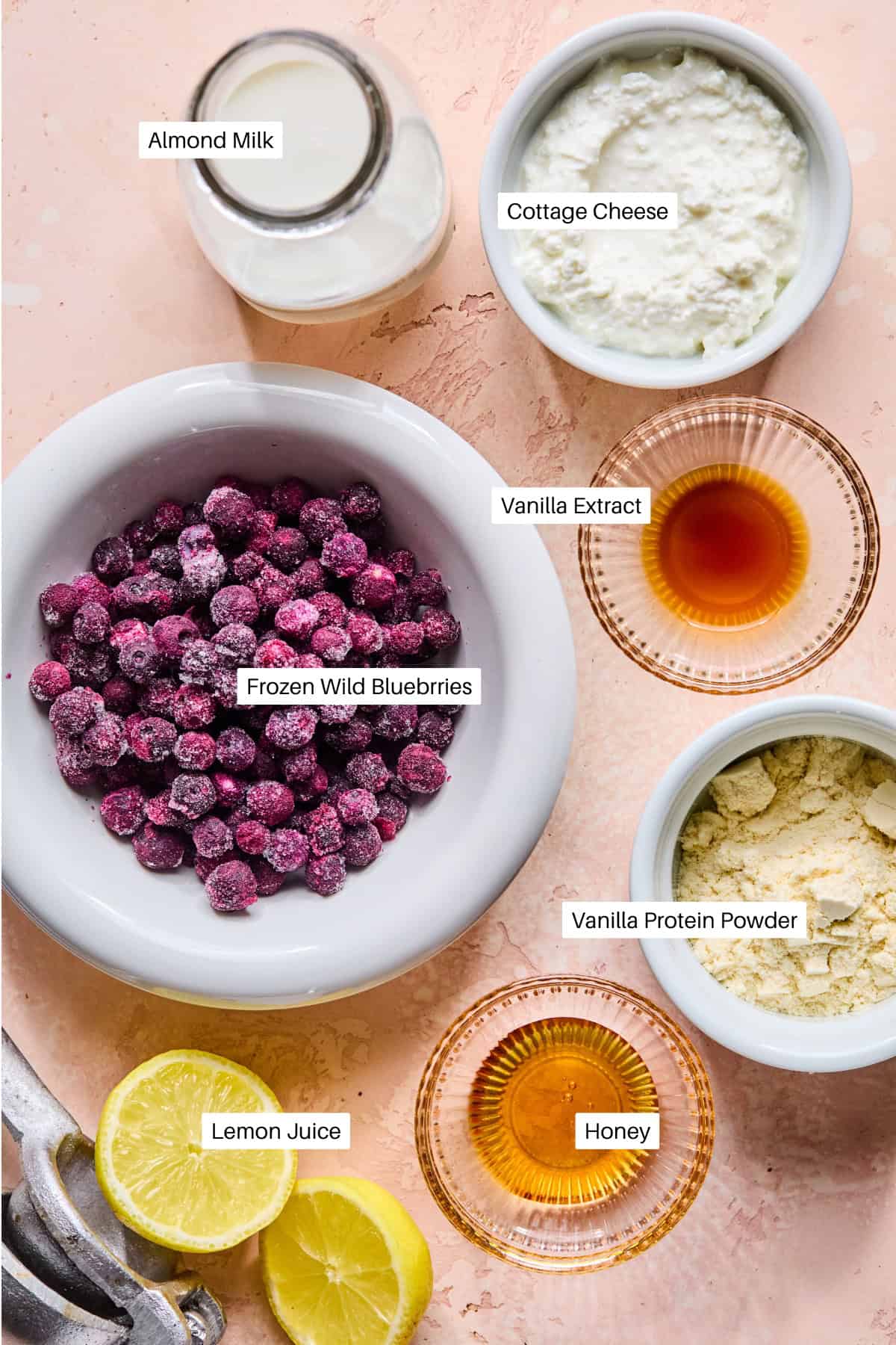 A flat lay of labeled ingredients for a blueberry protein smoothie in bowls on a peach surface: almond milk, cottage cheese, frozen wild blueberries, vanilla extract, vanilla protein powder, lemon juice, and honey. Sliced lemons are near the lemon juice.