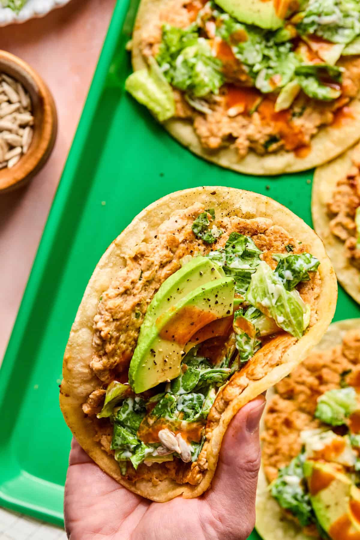 A hand holds a taco with refried beans, sliced avocado, lettuce, sunflower seeds, and hot sauce on a corn tortilla—no buffalo chicken smash tacos here, just vibrant veggie goodness. More similar tacos rest on a green tray in the background.