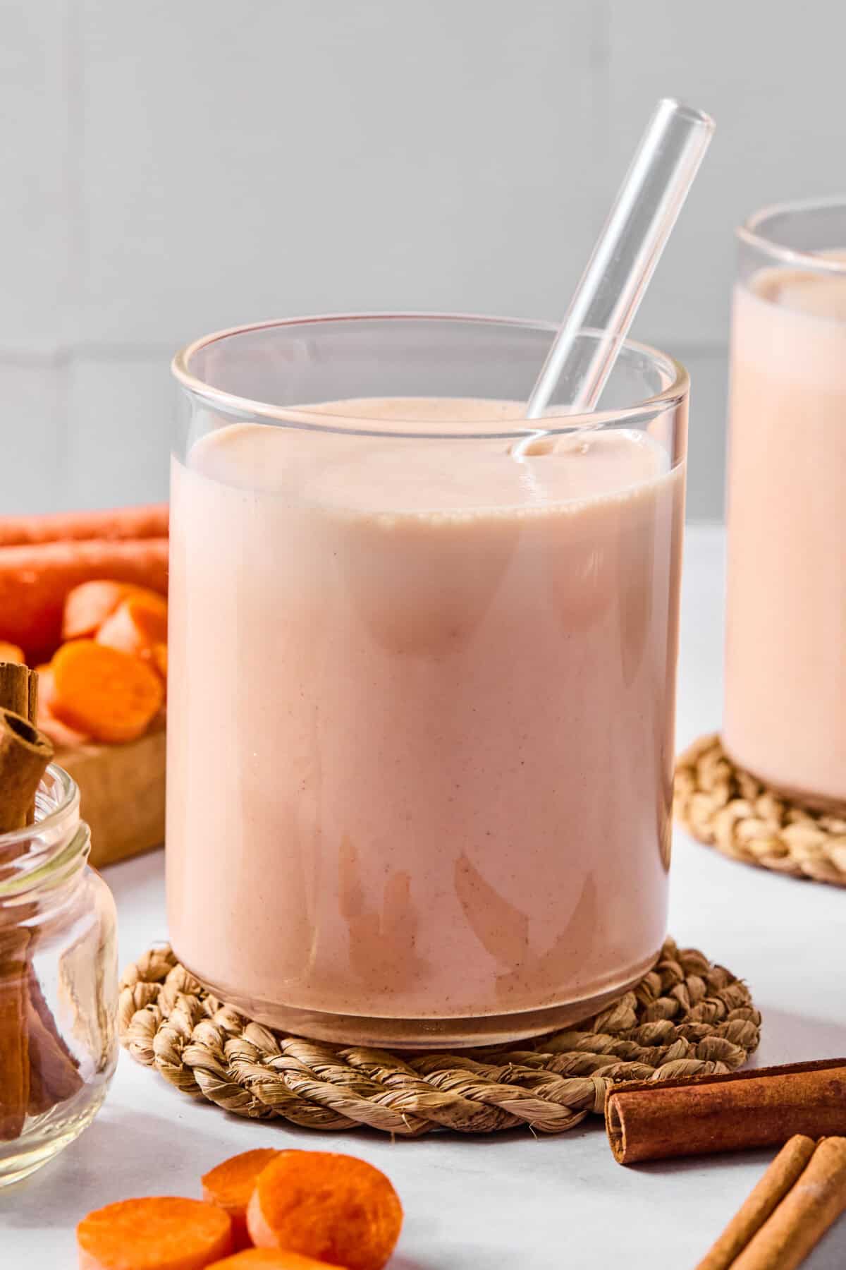 A glass of creamy carrot cake smoothie with a clear straw sits on a woven coaster, surrounded by cinnamon sticks, sliced carrots, and a small jar of cinnamon on a light surface.