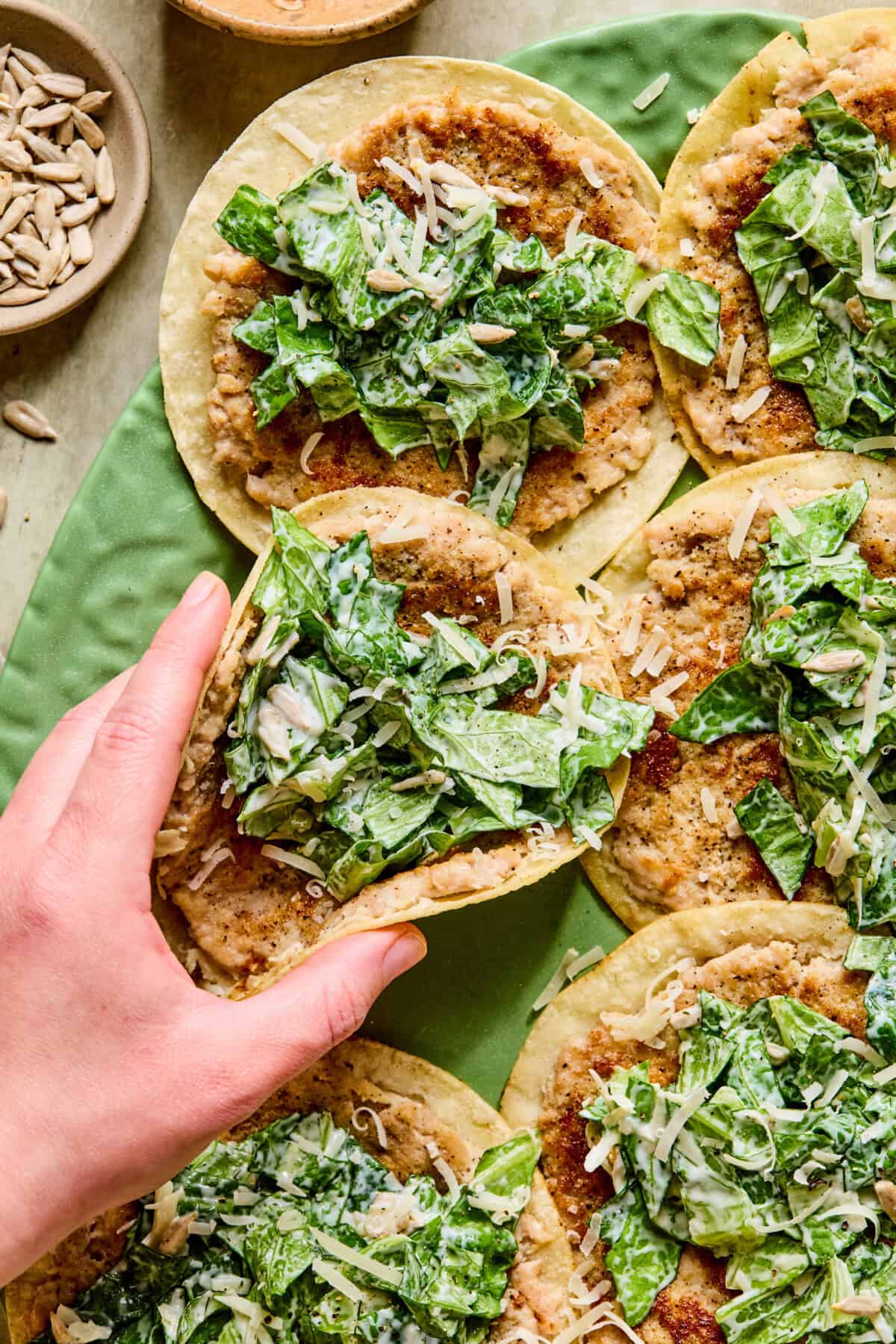 A hand picking up a smashed chicken caesar taco with a corn tortilla, creamy lettuce filling, shredded cheese, and a spread, surrounded by similar tacos on a green plate. A small bowl of sunflower seeds is visible in the corner.
