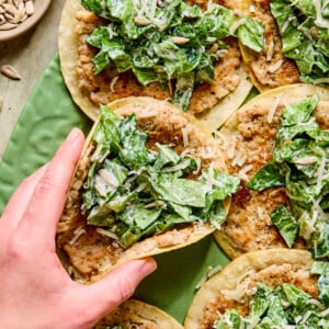 A hand picking up a smashed chicken caesar taco with a corn tortilla, creamy lettuce filling, shredded cheese, and a spread, surrounded by similar tacos on a green plate. A small bowl of sunflower seeds is visible in the corner.