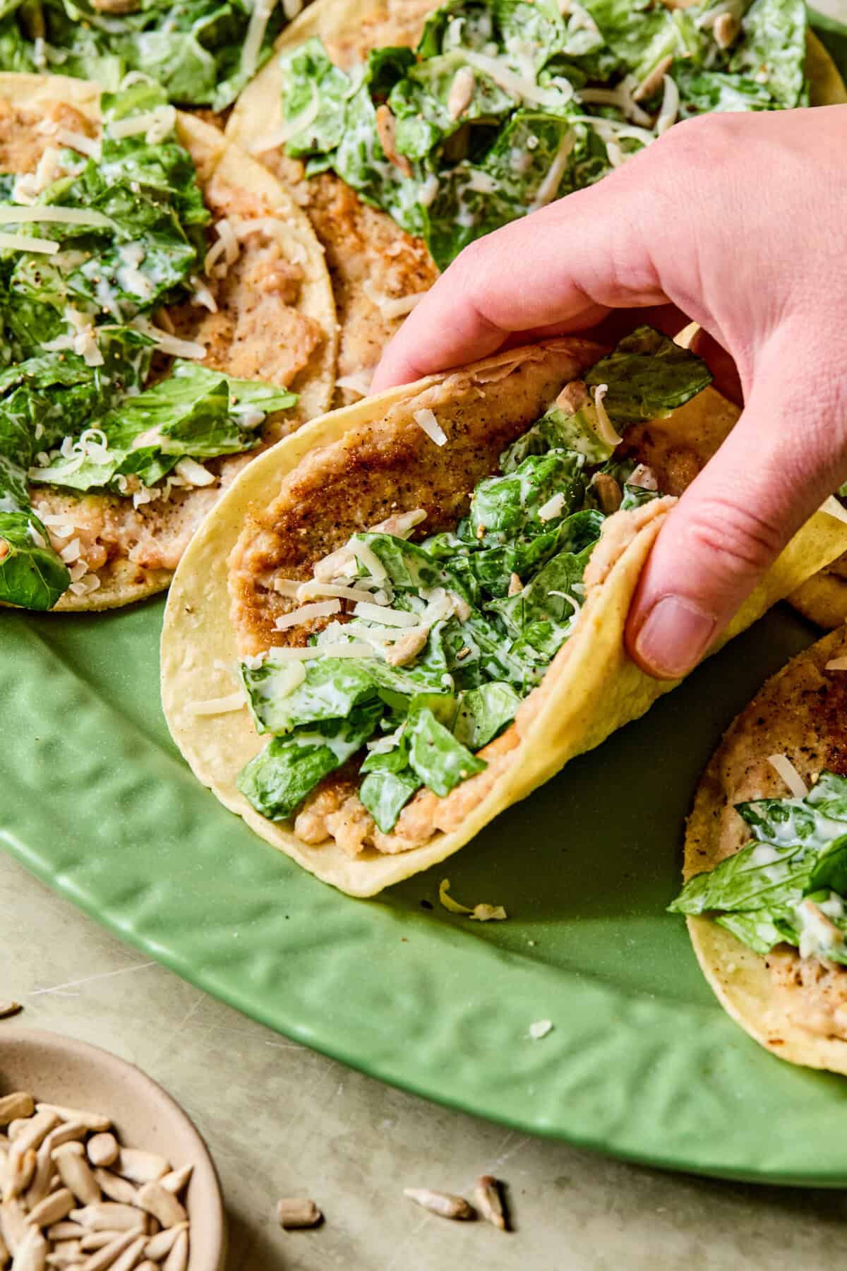 A hand picking up a smashed chicken caesar taco filled with refried beans, chopped romaine lettuce, shredded cheese, and creamy dressing from a green plate with several similar tacos.