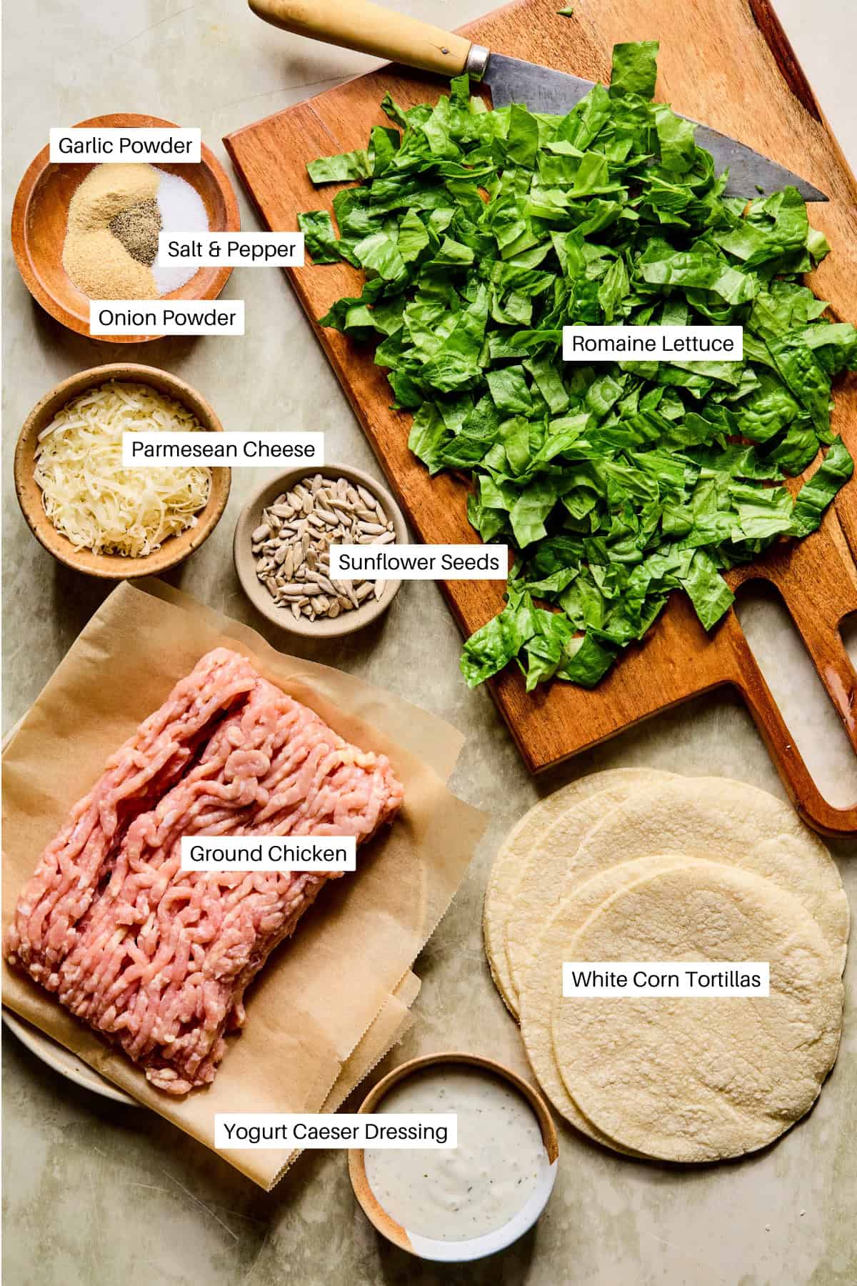 Chopped romaine lettuce on a cutting board, surrounded by labeled ingredients: garlic powder, salt & pepper, onion powder, Parmesan cheese, sunflower seeds, ground chicken, yogurt Caesar dressing, and white corn tortillas for smashed chicken caesar tacos.