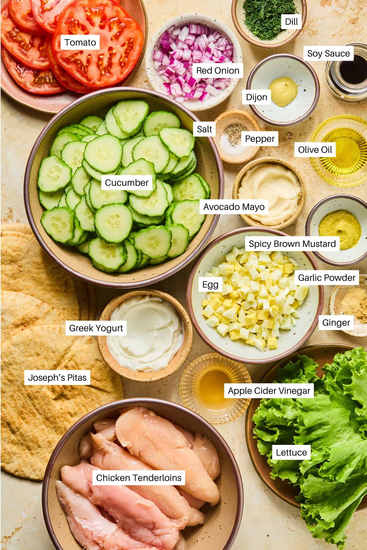 An overhead view of labeled ingredients for a delicious pita wrap recipe, including sliced tomato, cucumber, red onion, chicken tenderloins, lettuce, Greek yogurt, egg, pitas, and various seasonings and sauces in small bowls.