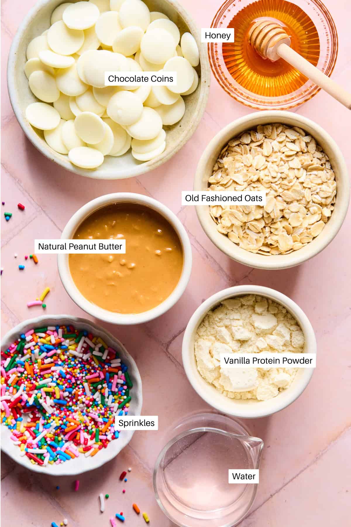 A vibrant spread of ingredients for protein balls on a pink surface, featuring white chocolate coins, honey, old fashioned oats, natural peanut butter, vanilla protein powder, colorful sprinkles, and a small pitcher of water.