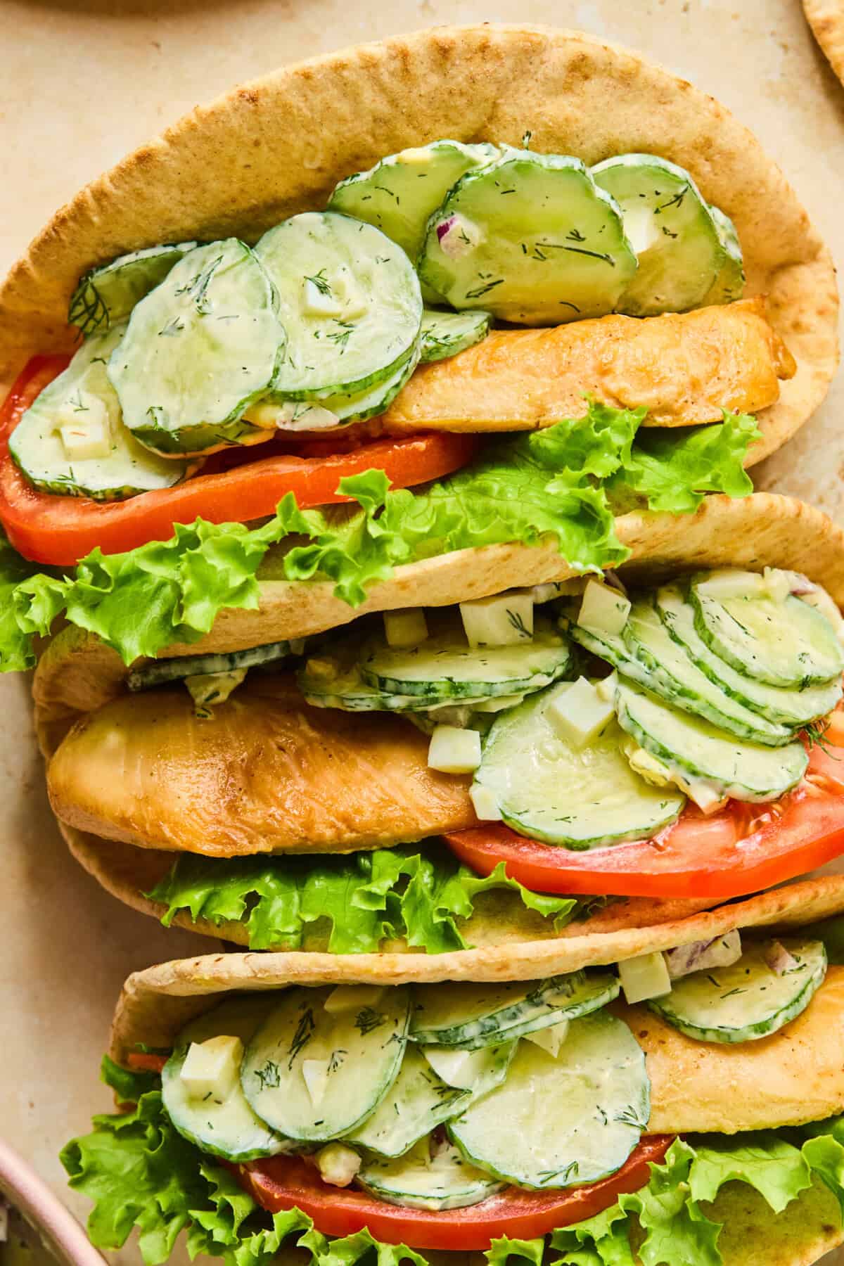 Three pita bread sandwiches filled with lettuce, tomato slices, grilled chicken, and a creamy cucumber salad with dill and cheese are arranged closely together on a light surface—a fresh take on any pita wrap recipe.