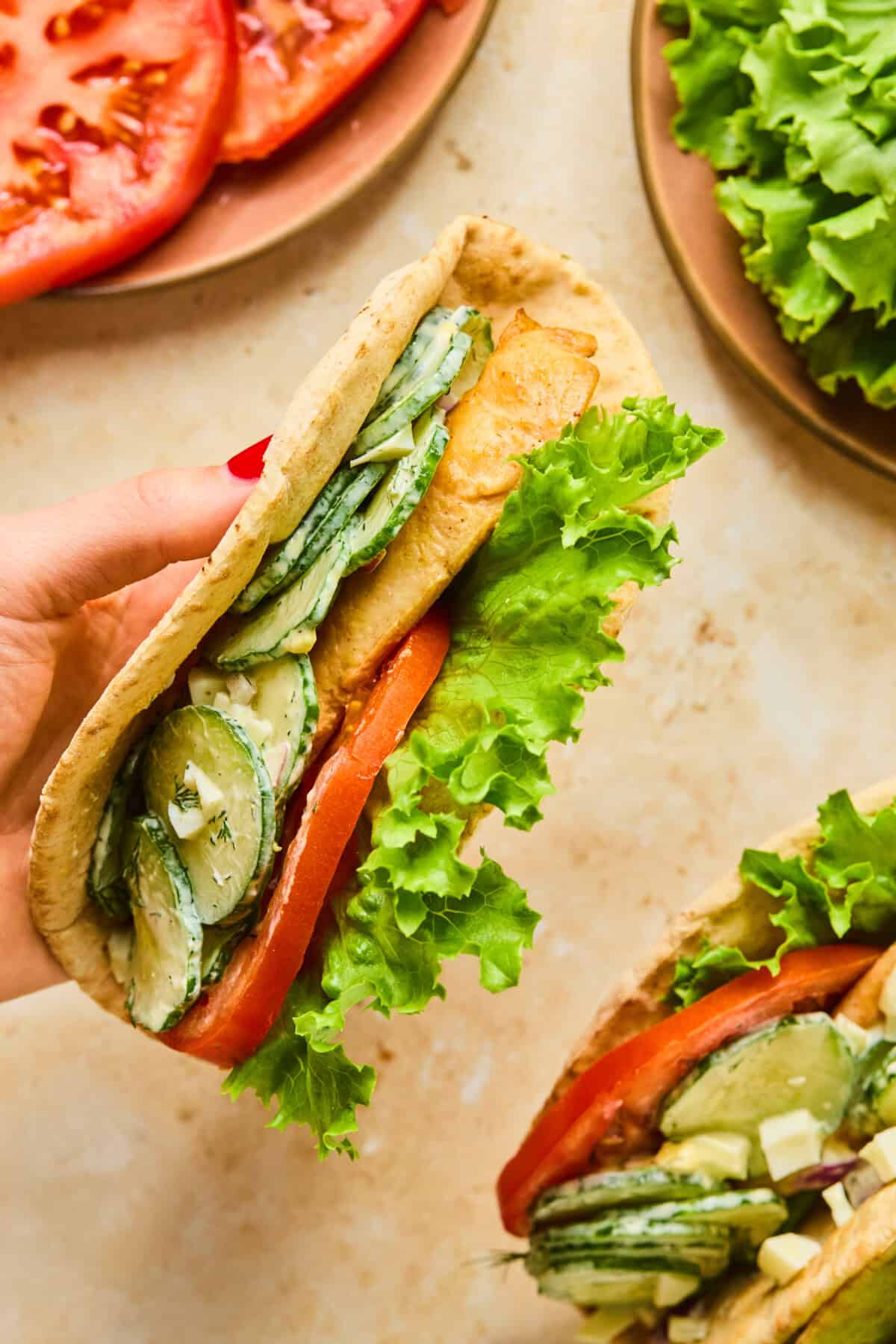 A hand holds a pita wrap stuffed with crispy tofu, fresh lettuce, tomato slices, cucumber, and creamy dressing. Plates with sliced tomatoes and lettuce are visible in the background—a delicious pita wrap recipe in action.