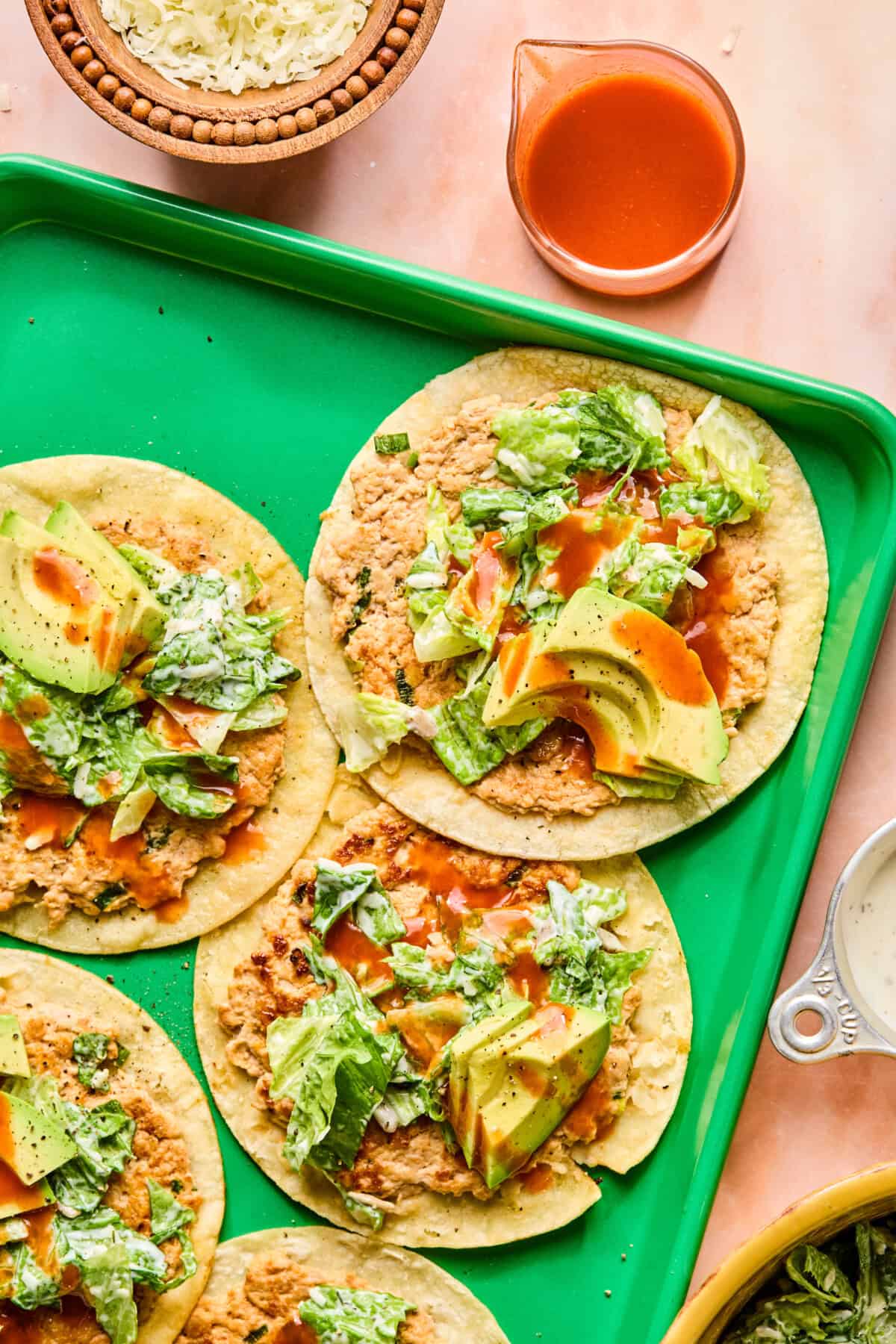 Four tostadas topped with refried beans, sliced avocado, lettuce, and hot sauce are arranged on a green tray—perfect alongside buffalo chicken smash tacos. A small bowl of shredded cheese and a cup of red sauce are nearby.