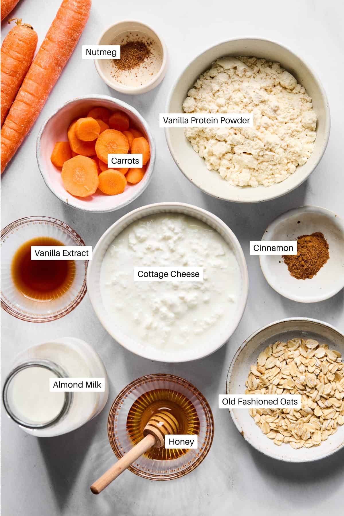 A flat lay of labeled ingredients in bowls for a carrot cake smoothie recipe, including carrots, nutmeg, vanilla protein powder, cinnamon, cottage cheese, vanilla extract, almond milk, honey, and old fashioned oats. Two whole carrots are shown.