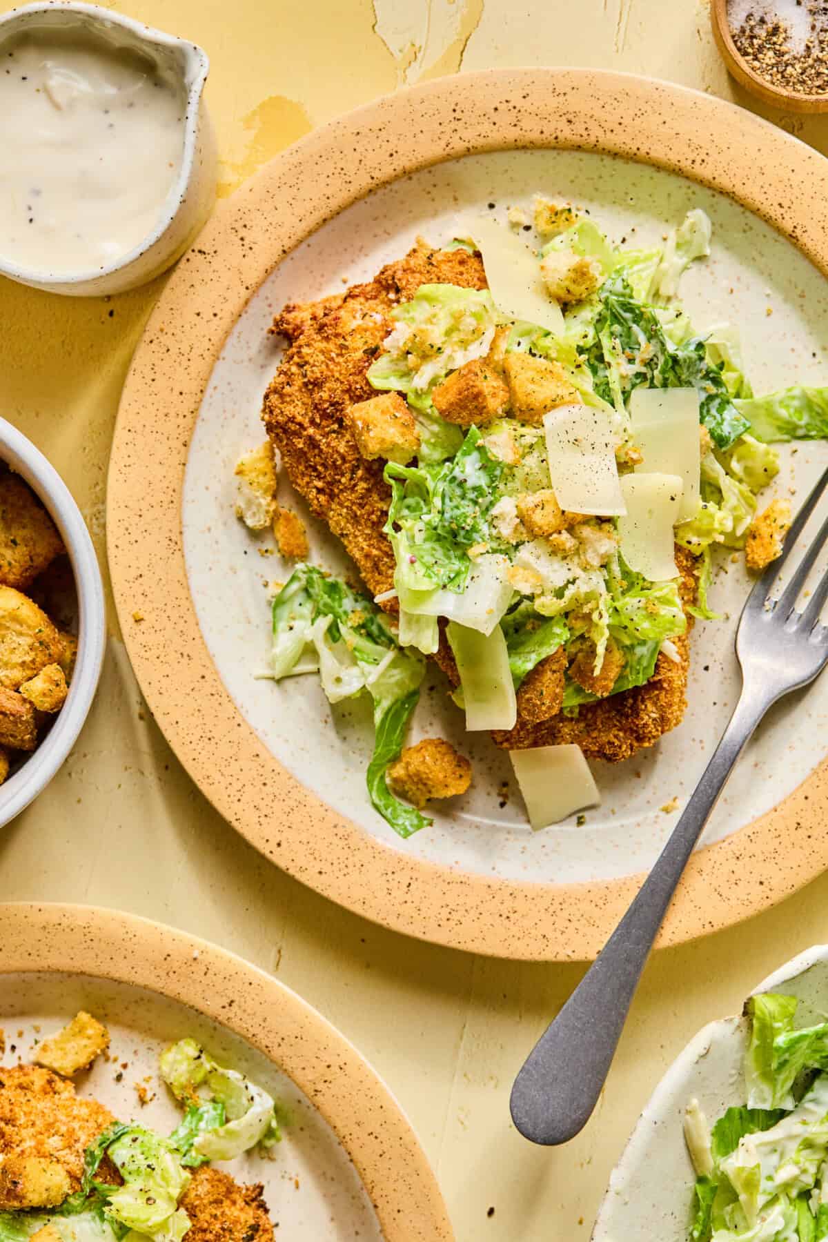 A plate of crispy chicken caesar cutlets topped with romaine lettuce, croutons, and shaved Parmesan cheese. A fork rests on the plate, and Caesar dressing is pictured nearby.