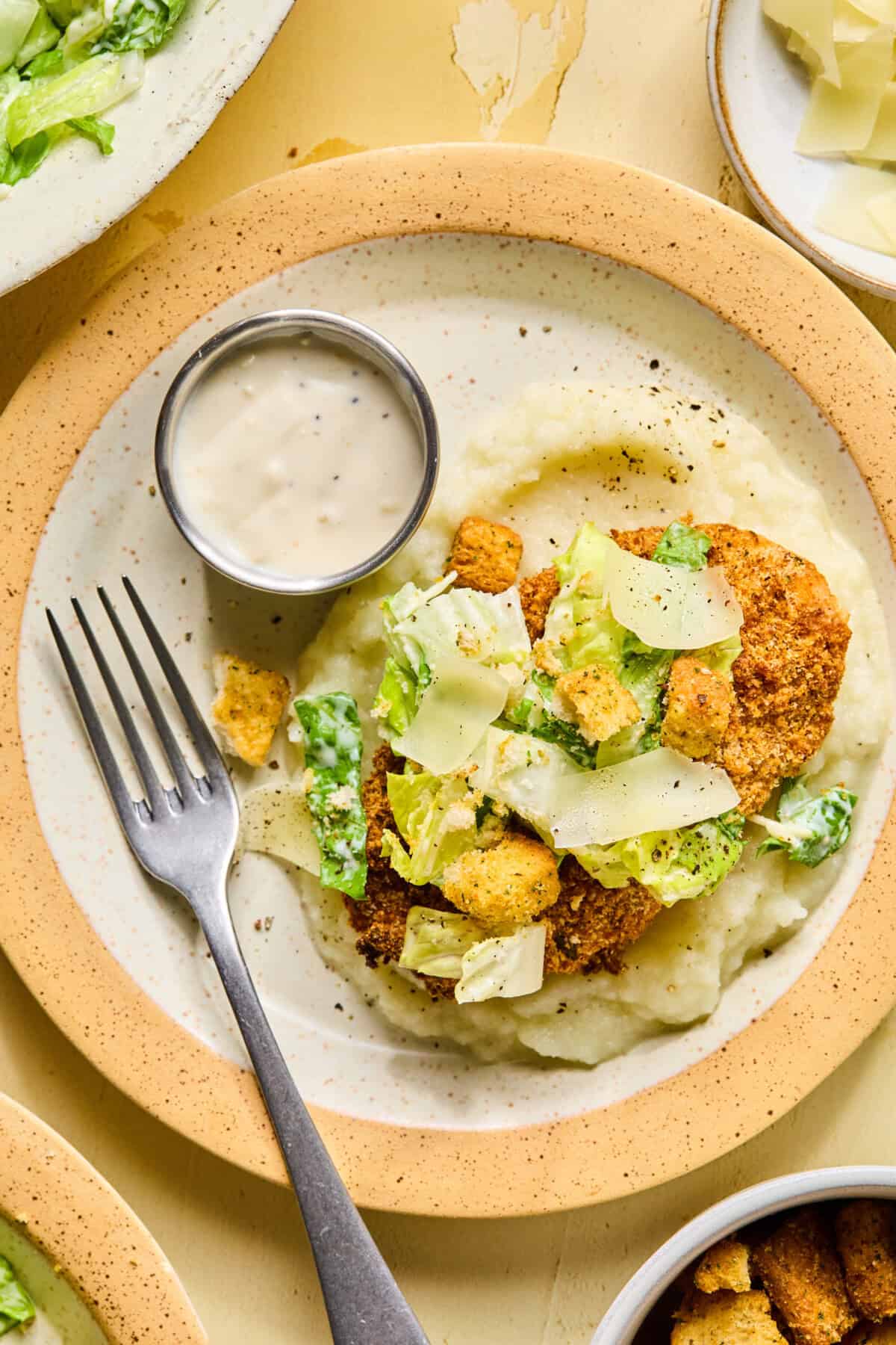 A plate featuring chicken caesar cutlets on mashed potatoes, topped with Caesar salad, croutons, and shaved cheese. A small cup of creamy dressing and a fork are on the side.