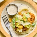 A plate featuring chicken caesar cutlets on mashed potatoes, topped with Caesar salad, croutons, and shaved cheese. A small cup of creamy dressing and a fork are on the side.