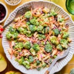 A large white bowl of Broccoli Crunch Salad with apples, shredded carrots, sunflower seeds, and dried cranberries is being tossed with serving utensils. Nearby are apples, sunflower seeds, and green glasses on a yellow surface.