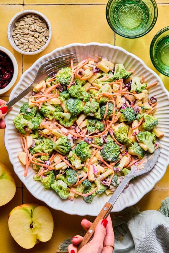 A large white bowl of Broccoli Crunch Salad with apples, shredded carrots, sunflower seeds, and dried cranberries is being tossed with serving utensils. Nearby are apples, sunflower seeds, and green glasses on a yellow surface.