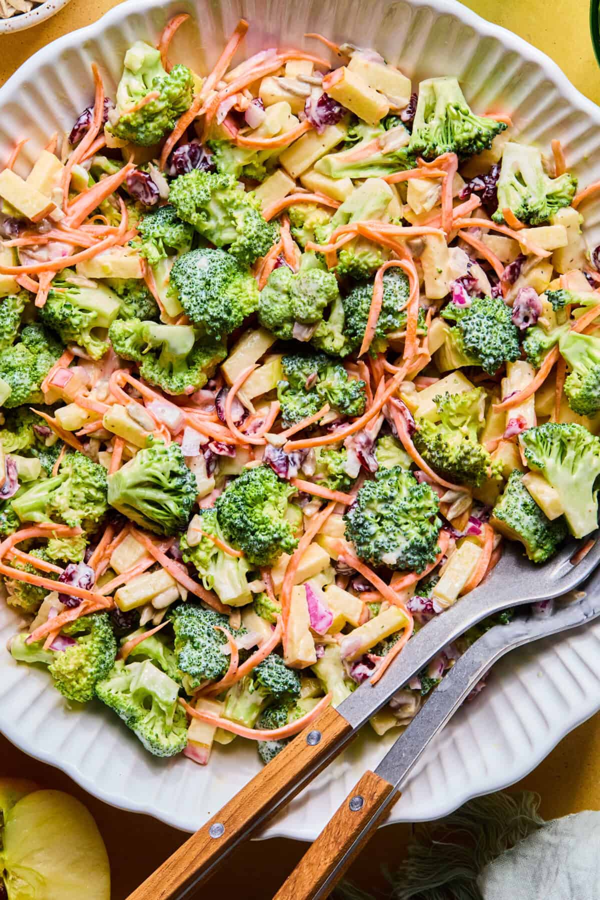 A close-up of Broccoli Crunch Salad in a white dish, featuring broccoli florets, shredded carrots, chopped apples, raisins, and a creamy dressing. Two wooden-handled serving utensils rest in the salad.