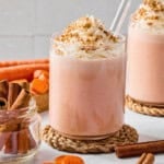 Two glasses of creamy carrot cake smoothie topped with whipped cream and crushed nuts, each with a clear straw. Surrounded by cinnamon sticks, carrot slices, and whole carrots on a light surface.