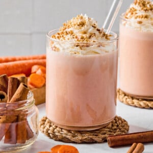 Two glasses of creamy carrot cake smoothie topped with whipped cream and crushed nuts, each with a clear straw. Surrounded by cinnamon sticks, carrot slices, and whole carrots on a light surface.