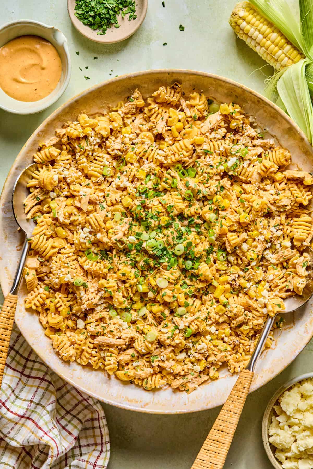 A large dish of Mexican street corn pasta salad mixed with green onions and a creamy orange-colored sauce, garnished with chopped herbs. Slices of corn, sauce, and chopped herbs are placed around the main dish.