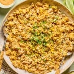 A large dish of Mexican street corn pasta salad mixed with green onions and a creamy orange-colored sauce, garnished with chopped herbs. Slices of corn, sauce, and chopped herbs are placed around the main dish.